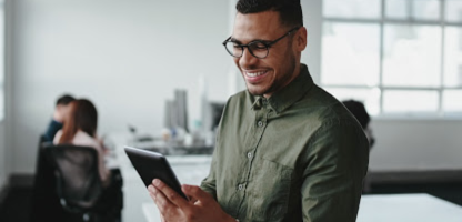 Man wearing glasses and a green shirt smiling while using a tablet in a modern office; two people are blurred in the background at a desk