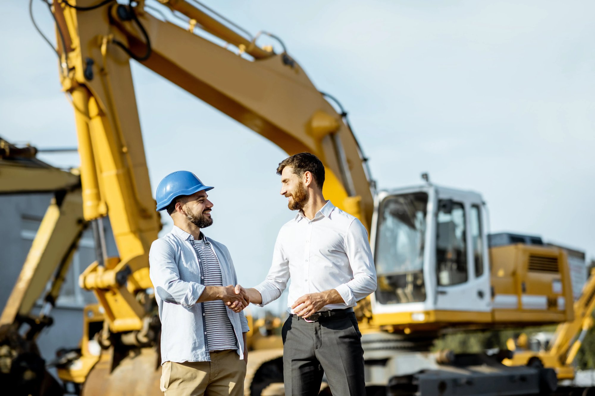 Business professional and construction worker shaking hands in front of construction equipment at a job site
