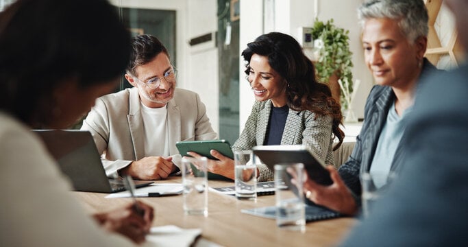 Three professionals sitting at a conference table during a business meeting, reviewing work on tablets and a laptop