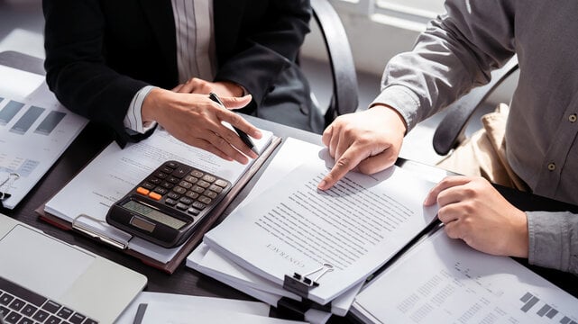 Two male professionals reviewing paperwork on a desk next to a calculator and laptop