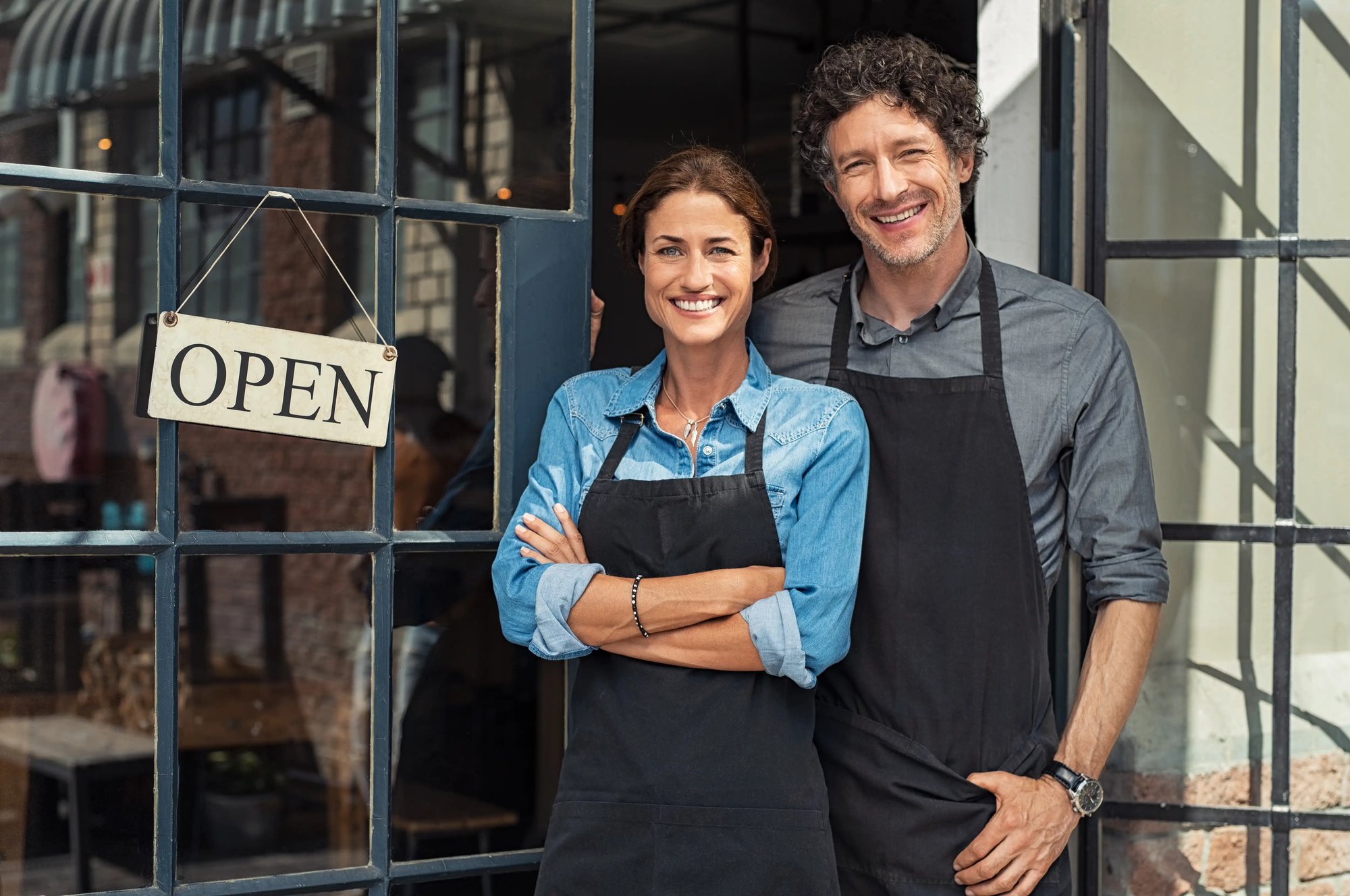 Two small business owners standing outside their storefront wearing work aprons and smiling