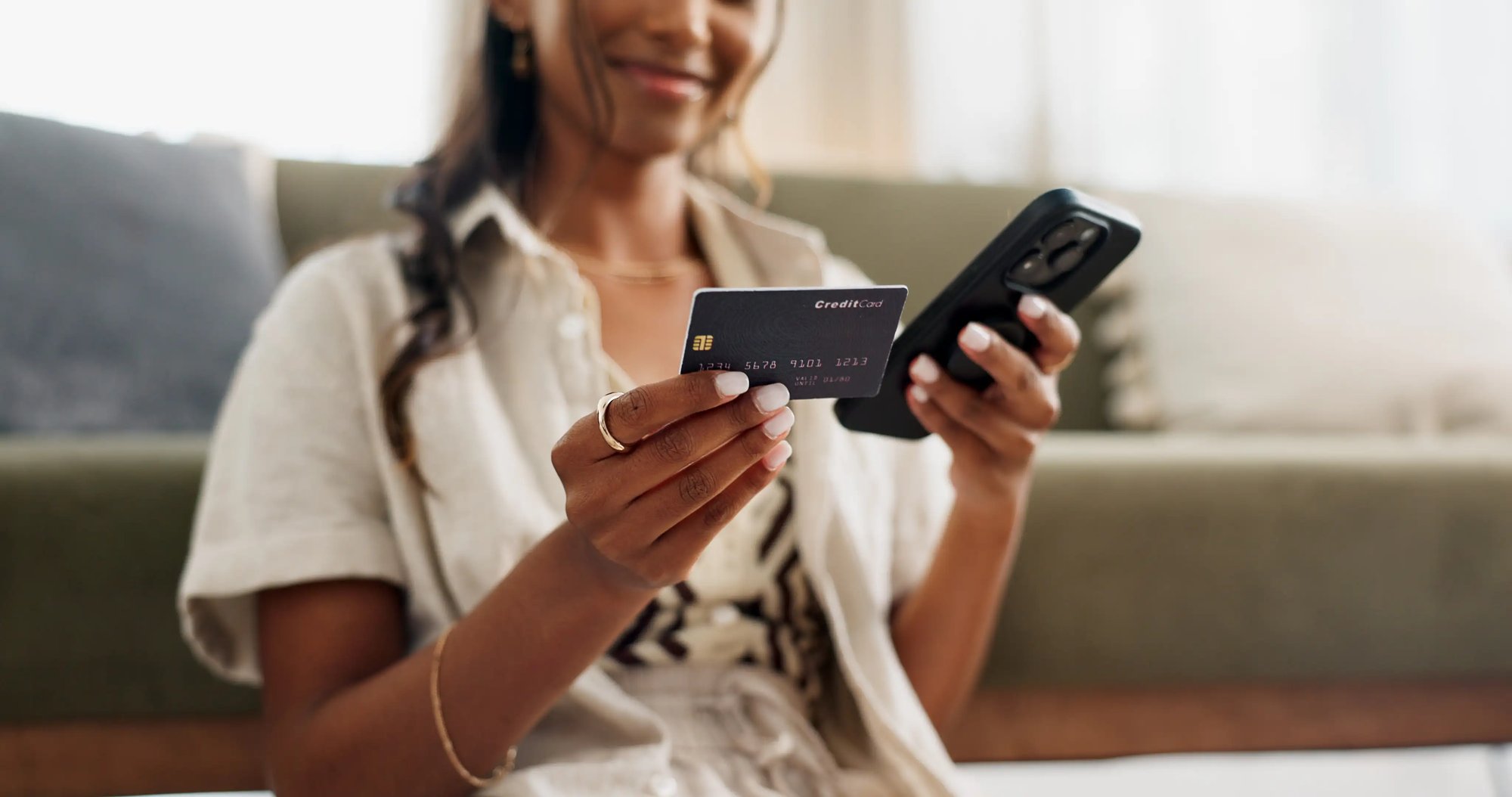 Woman sitting on the floor in front of her couch, holding her smartphone in one hand and payment card in the other