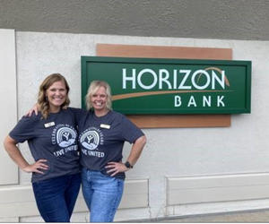 Two female Horizon Bank employees stand in front of a Horizon Bank sign wearing United Way shirts.