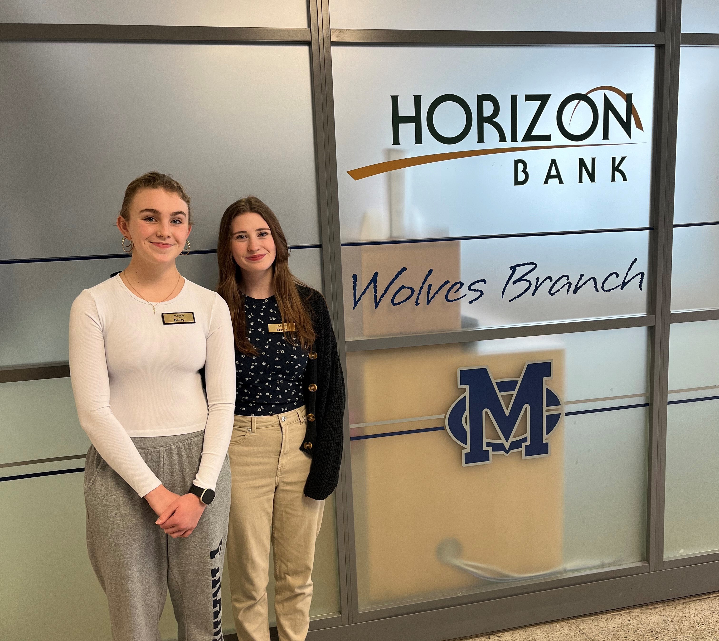 Two female high school students stand in front of the Horizon Bank Wolves branch.